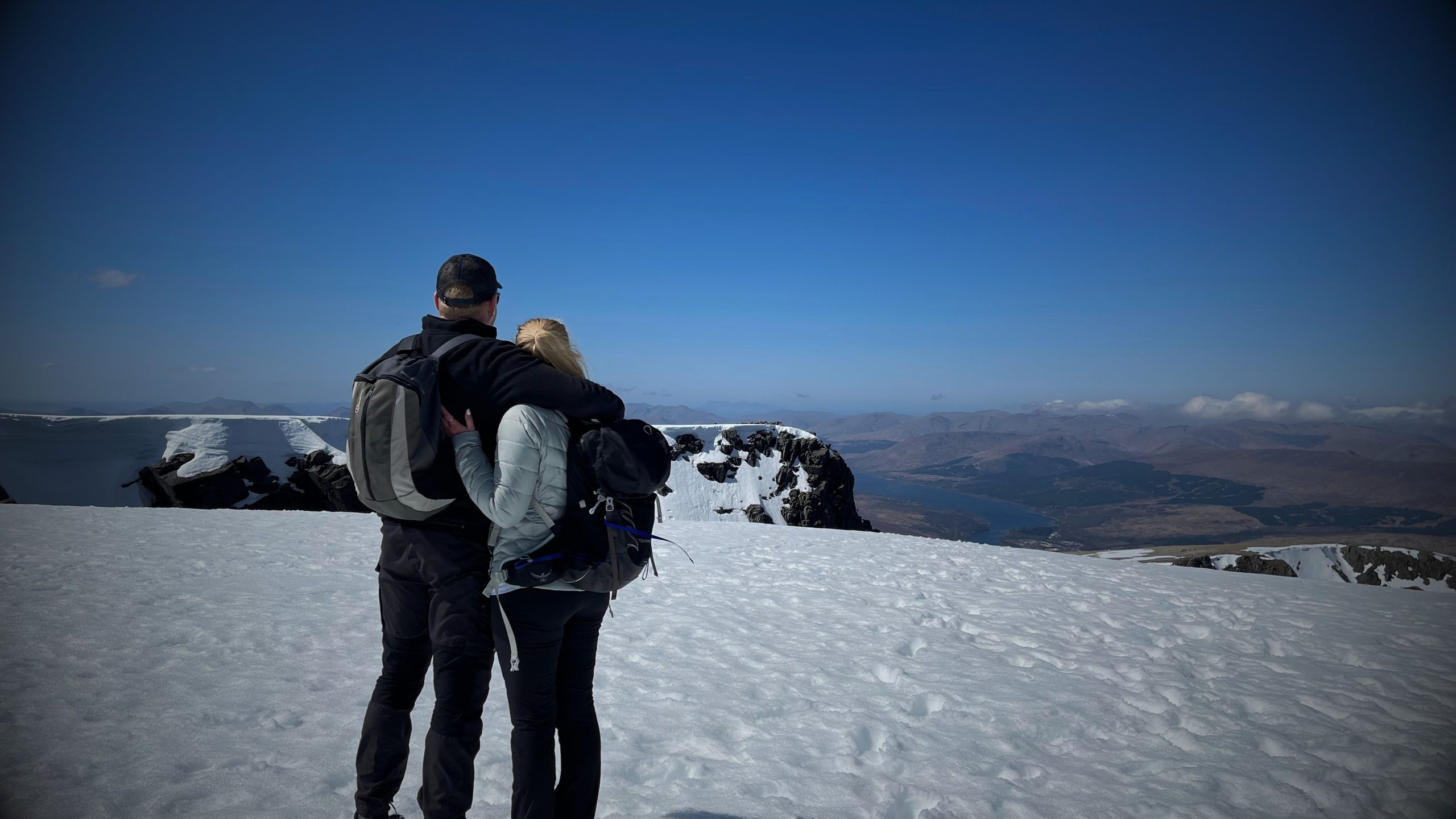 James Masters and his wife in winter hiking gear standing on a snow-covered mountain summit, arm-in-arm, looking out at a wide mountainous landscape under a clear blue sky.