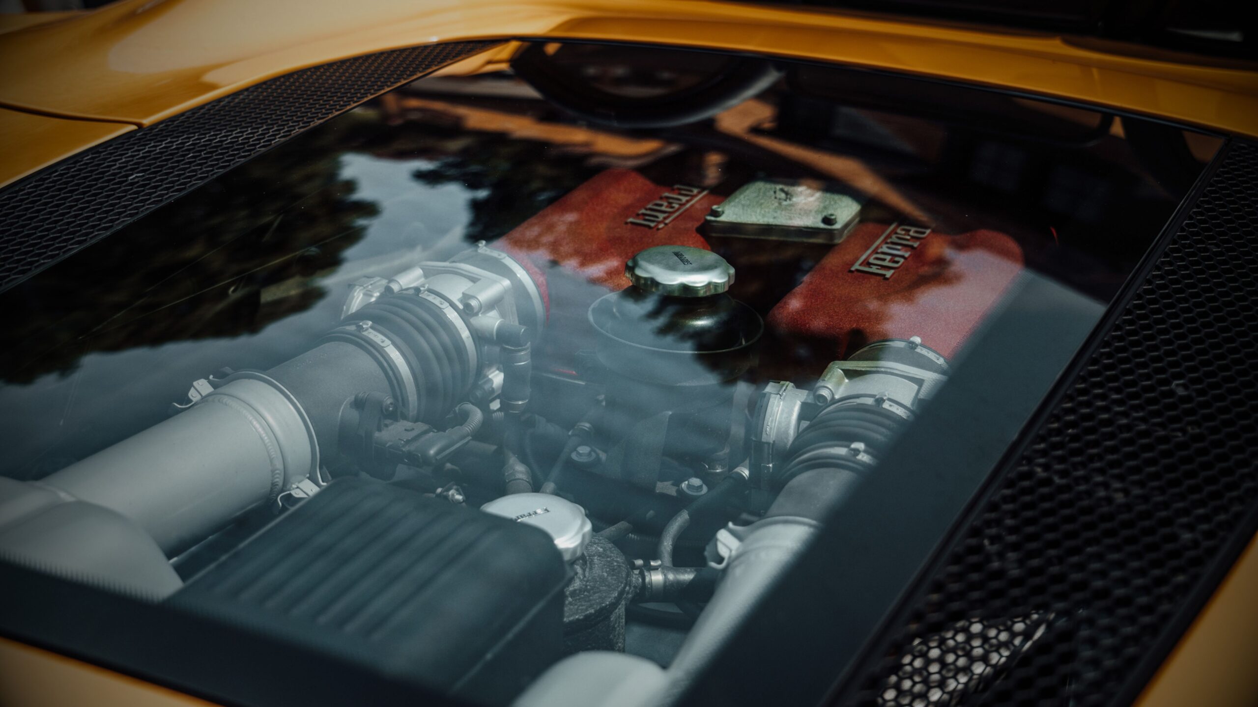 Ferrari Engine Glass Cover Close-up view through the clear rear glass engine cover of a yellow Ferrari, displaying the mid-mounted engine bay with red intake manifolds.