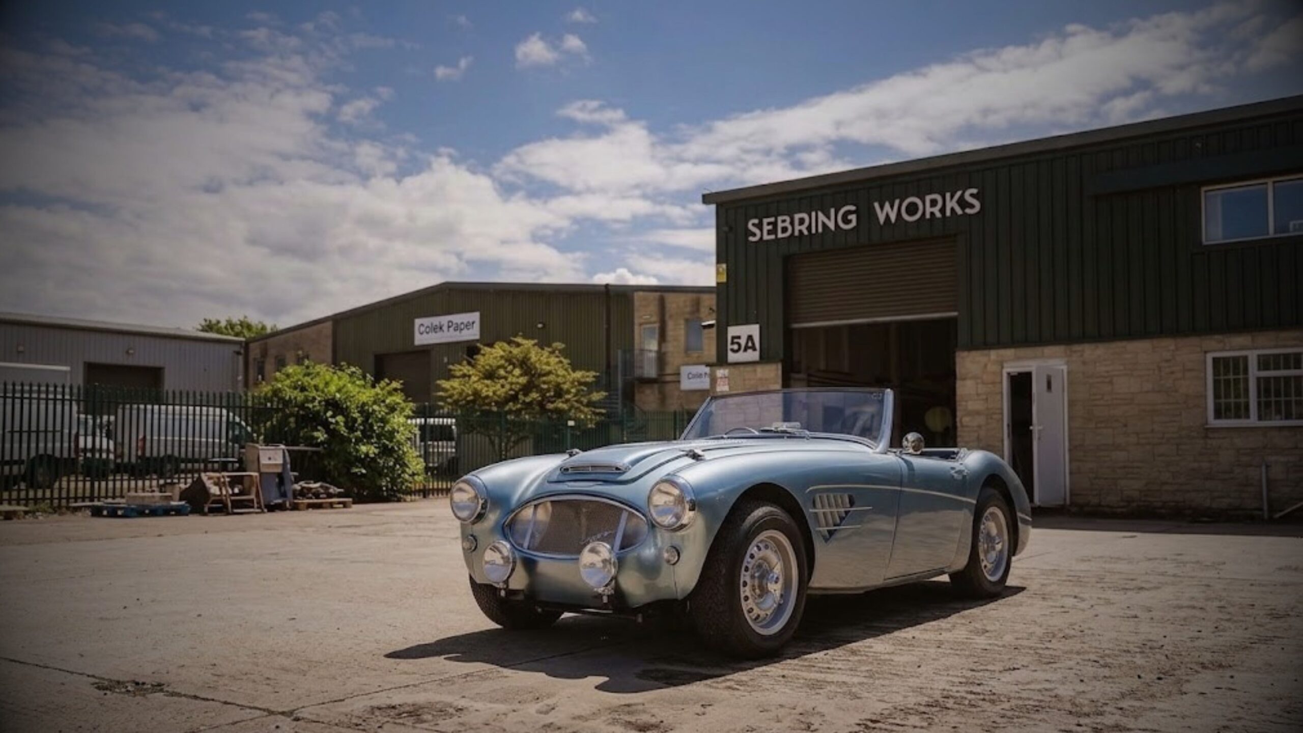 Classic light blue Sebring convertible sports car parked on an open concrete yard outside the Sebring Works workshop under a blue sky.